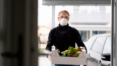 Homem usando máscara carrega uma caixa de entregas cheia de verduras.