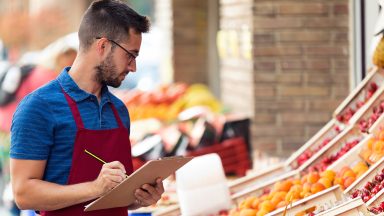 Homem de camisa polo azul e avental bordô segurando uma prancheta em frente a seção de verduras em um supermercado