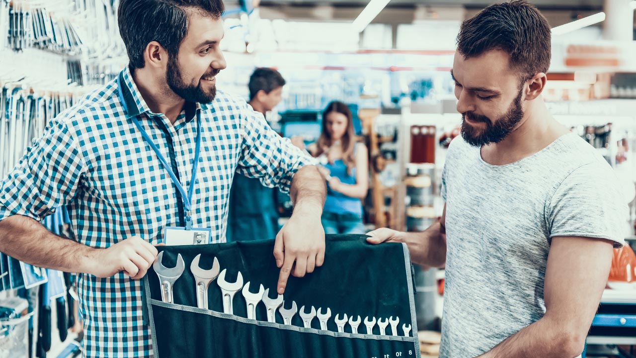 Dois homens em uma loja de ferragens discutem um rolo de chaves inglesas. Um homem aponta para as chaves inglesas enquanto o outro escuta. Prateleiras com ferramentas são visíveis ao fundo