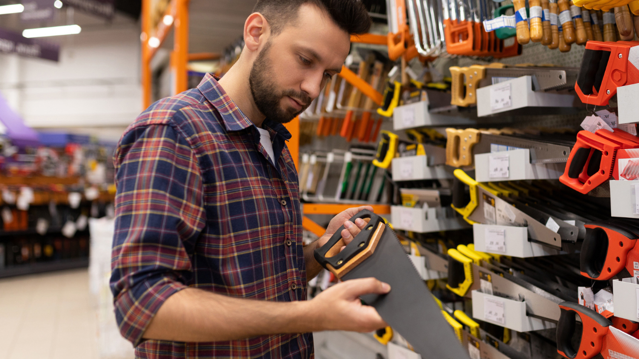 Homem segurando e olhando um serrote em uma loja de materiais de construção, com outras opções no paredão, representando o mix de produtos para materiais de construção.