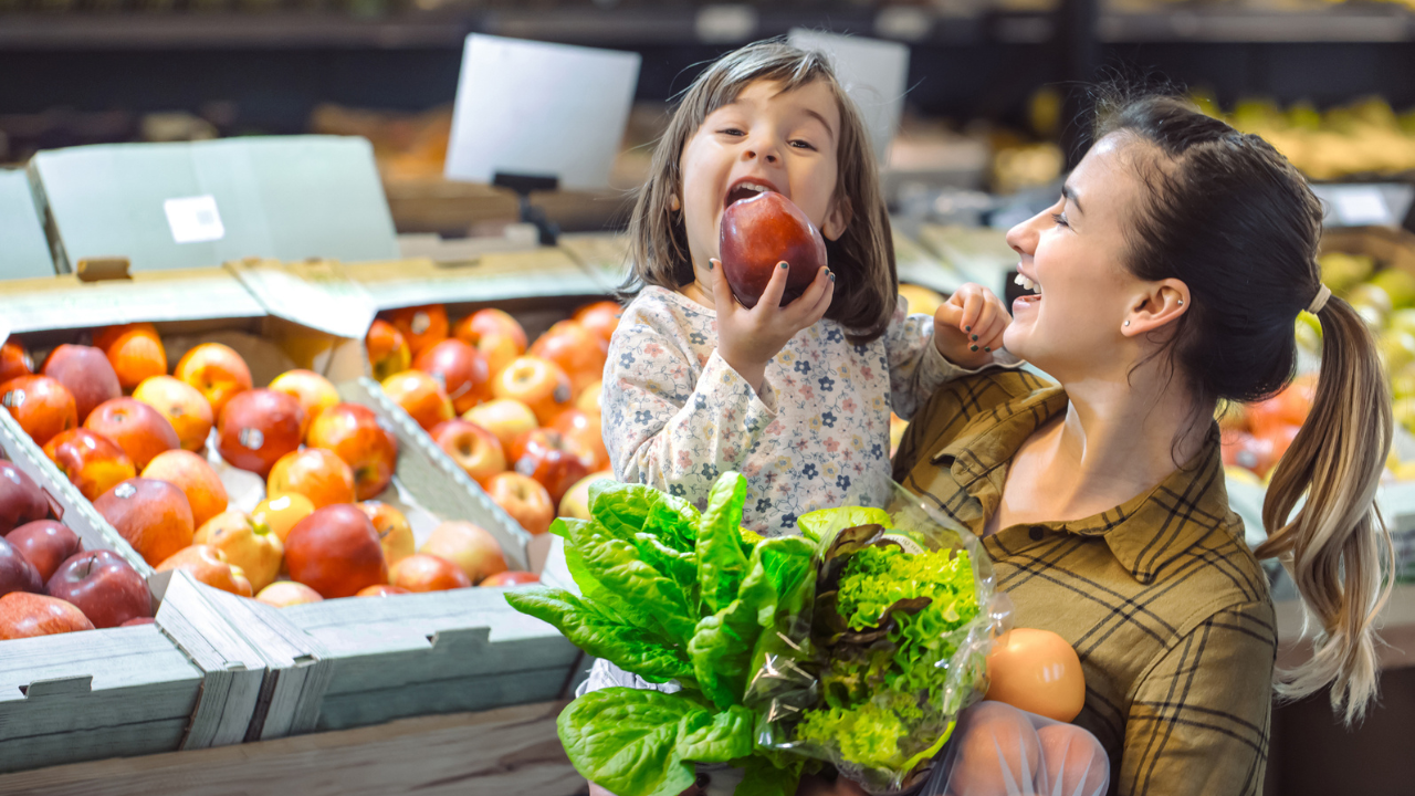Mãe e filha sorrindo enquanto fazem compras em uma feira, escolhendo frutas e verduras frescas para uma alimentação saudável.