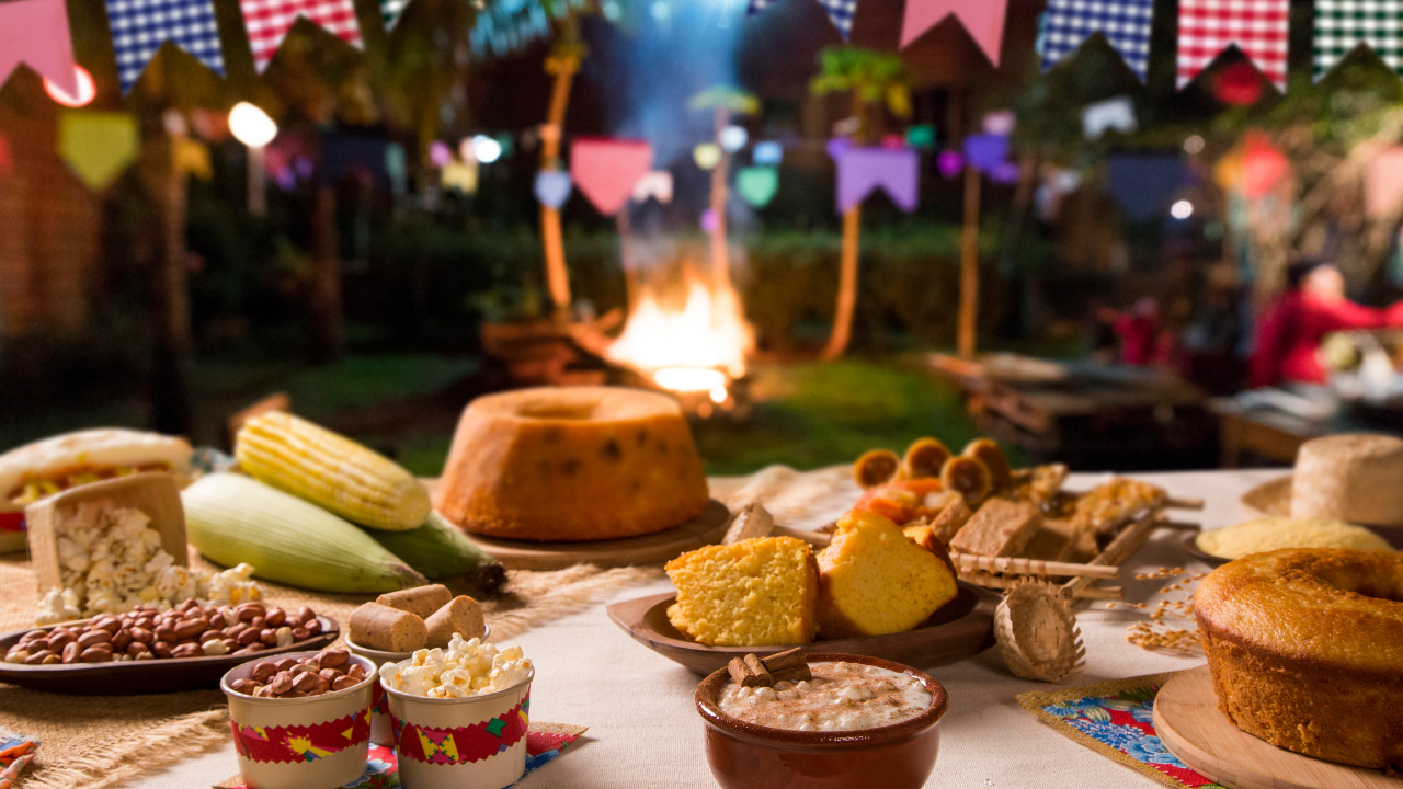 Mesa decorada com comidas típicas de festa junina, como canjica, milho, bolo de fubá, pipoca e amendoim, em um ambiente ao ar livre com bandeirinhas coloridas e fogueira ao fundo — representando a atmosfera tradicional do São João nos supermercados.