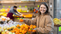 Mulher sorridente escolhe laranjas frescas em um supermercado, vestindo roupa de frio típica do inverno no Brasil, com frutas coloridas ao fundo e clima ameno característico da estação.