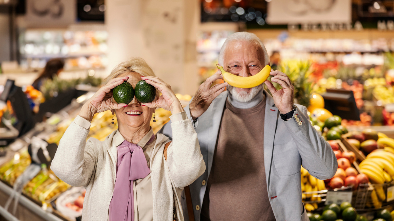 Casal idoso feliz se diverte com frutas no supermercado, aproveitando os benefícios de um programa de fidelidade.