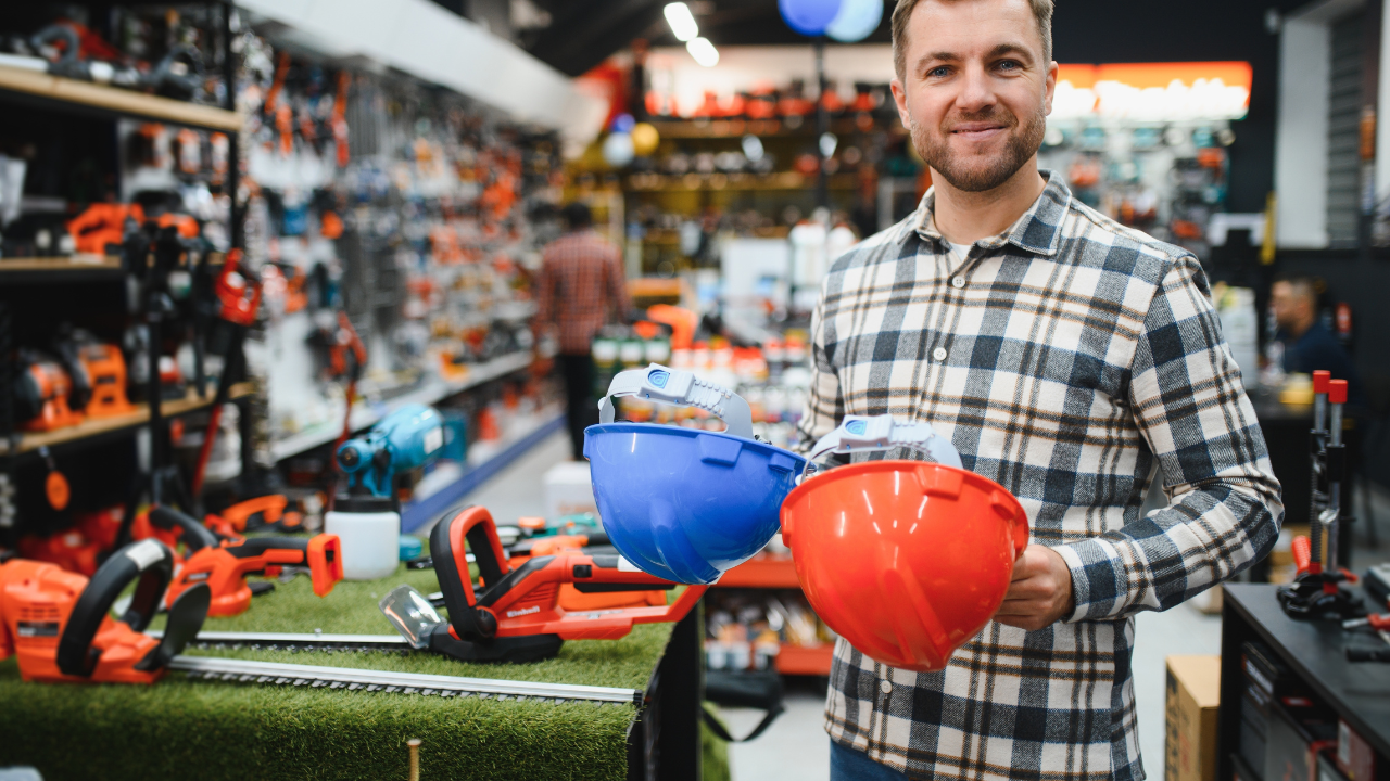 Empresário sorridente segurando capacetes de segurança azul e vermelho em loja repleta de ferramentas e equipamentos, ilustrando um case de sucesso no segmento de materiais de construção.