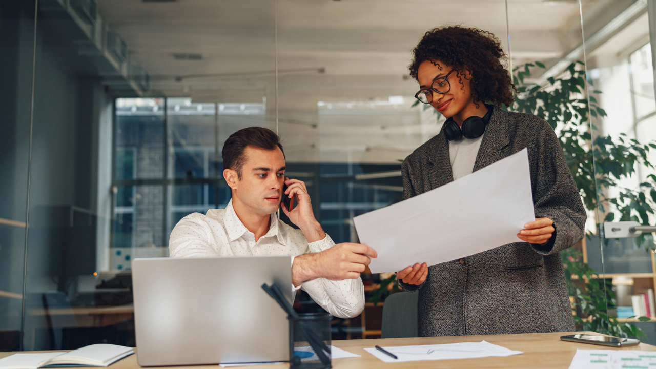 homem de negócios sentado em frente a um computador recebendo uma papel A3 das mãos de uma profissional com fones de ouvido. Ambos em um escritório moderno, representando o setor administrativo de um empreendimento varejos e simbolizando a necessidade de organização que se fará necessária com a nova reforma tributária no varejo.