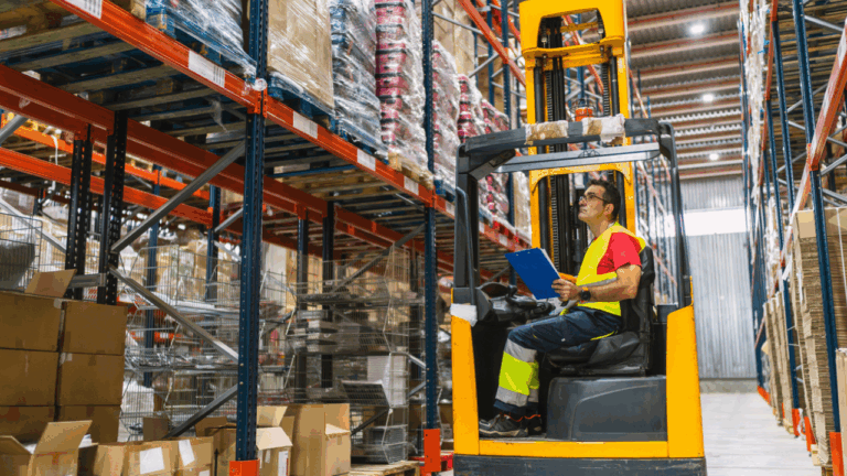 homem segurabdo uma prancheta azul enquanto opera uma máquina empilhadeira em um grande armazem com prateleiras altes, características centrais de grandes operações varejistas, indicando a necessidade de eficiência operacional nesse segmento,