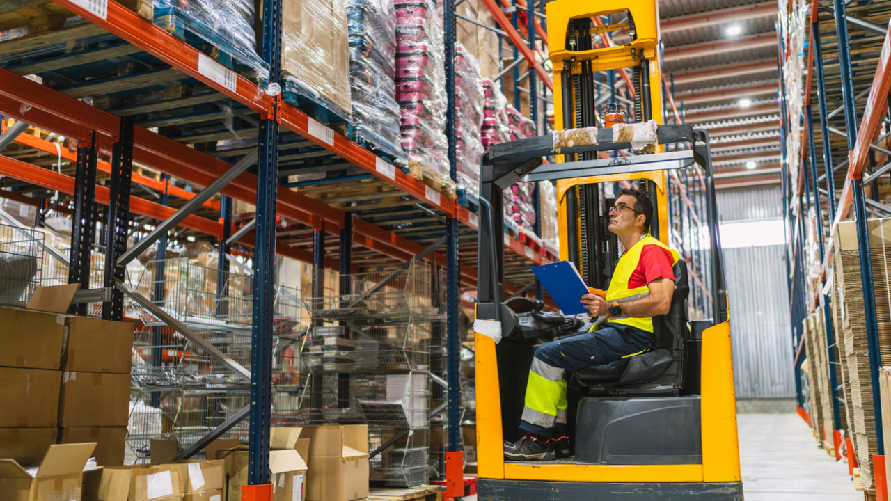 homem segurabdo uma prancheta azul enquanto opera uma máquina empilhadeira em um grande armazem com prateleiras altes, características centrais de grandes operações varejistas, indicando a necessidade de eficiência operacional nesse segmento,