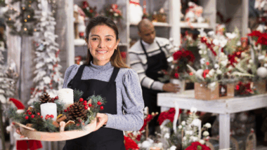 mulher em pé, na frente de uma bancada olhando para frente com uma cesta de enfeites de natal nas mãos com várias mercadorias em desfoque atrás, representando o período mais crítico do ano, o final de ano.