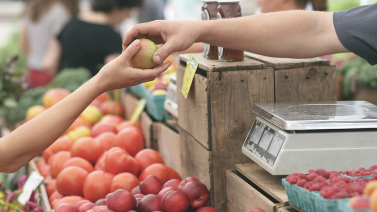 uma mão entregando uma maçã para outra pessoa com fundo de um supermercado na área de hortifruti representando a sustentabilidade no varejo
