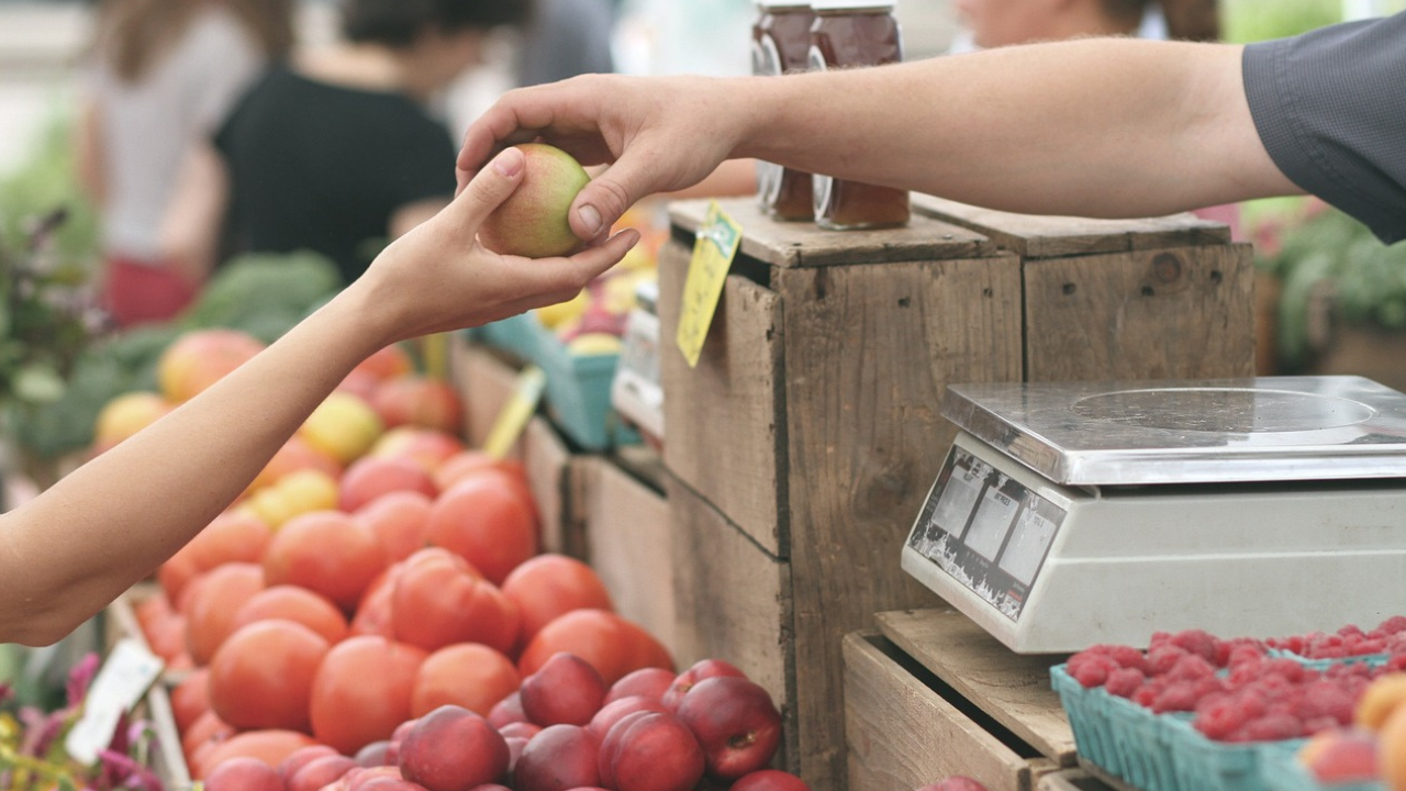 uma mão entregando uma maçã para outra pessoa com fundo de um supermercado na área de hortifruti representando a sustentabilidade no varejo